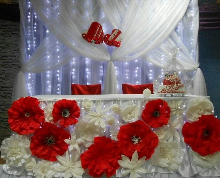 Table d'honneur de mariage décorée de grandes fleurs en papier rouges et blanches avec drapés rétroéclairés